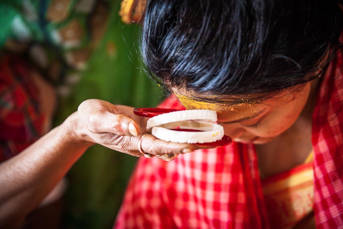 A Bengali bride, turmeric on her forehead, touches a pair of white conch shell sakha and red pola to her brow before they are put on